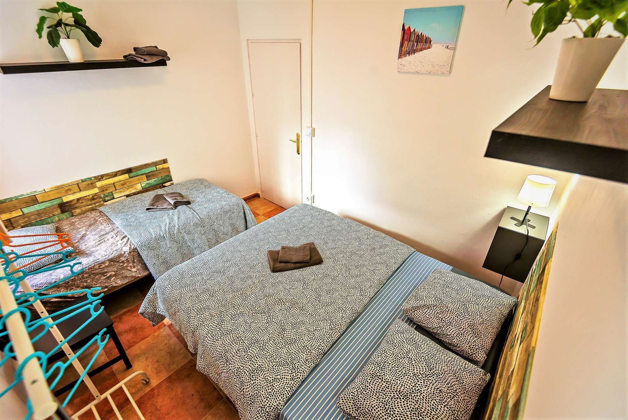 A small, well-lit room with two beds, a chair, a shelf with plants, and a door.