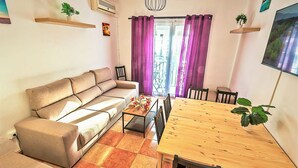 A living room with a sofa, a dining table with chairs, a small coffee table, and a view through the window. - Plaza Polvorista 4-Bedroom Apartment with Balcony in front of the beautiful park (El Puerto de Santa María)
