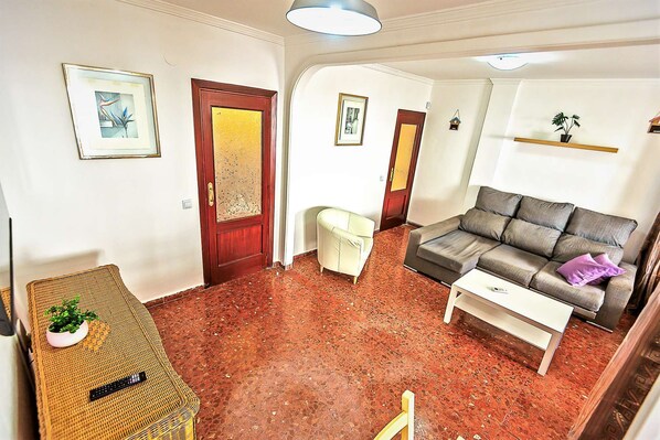 Una sala de estar con un sofá, una mesita, un sillón de mimbre y piso cubierto de alfombra roja.