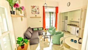 Una sala moderna con un sofá, sillas verdes, una mesa de comedor con tazas, una cocina con armarios y electrodomésticos, y un ventanal con cortinas.