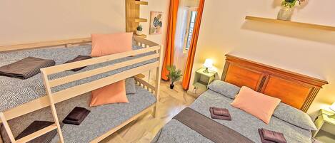 A room with bunk beds, a bedside table with a lamp, a small shelf with flowers, and a window with curtains.