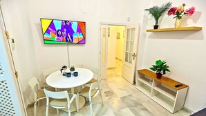 Una sala moderna con una mesa de comedor redonda, sillas blancas, un televisor de pantalla plana montado en la pared y un estante de madera con plantas.
