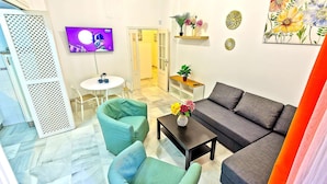 Una sala de estar moderna con un sofá gris, sillones color turquesa, una mesa de centro y un televisor en la pared. Se observan platos decorativos en la pared y una planta en una estantería.