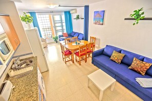 Ein modernes Wohnzimmer mit einem blauen Sofa, einem Esstisch und Stühlen. Es gibt eine Küchenzeile mit einer Mikrowelle und einem Spülbecken.