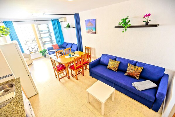 Ein Wohnzimmer mit blauem Sofa, einem Esstisch mit Stühlen, einem kleinen weißen Tisch und einer Küchenzeile mit Kühlschrank.