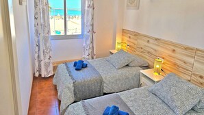 A room with two beds, a window with a view of the beach, a wooden headboard, and a shelf with decorative items.