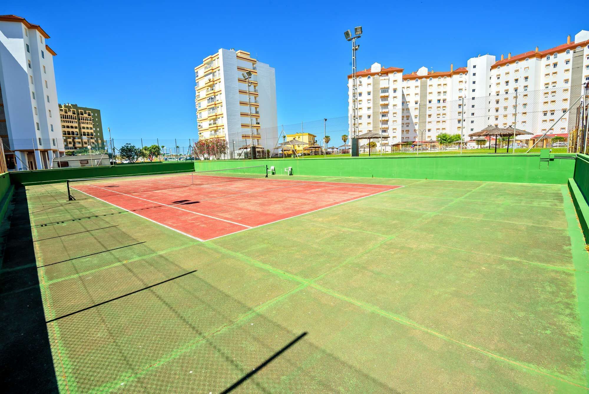 A tennis court with a net, surrounded by green fencing and buildings in the background.
