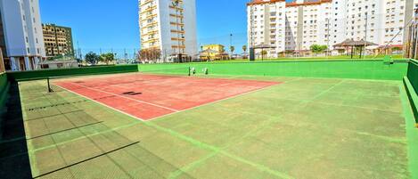 A tennis court with a net, surrounded by green fencing and buildings in the background.