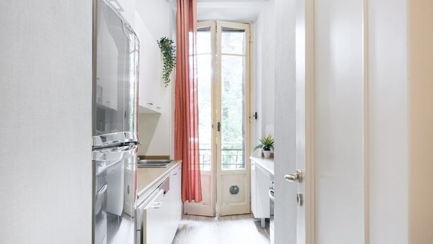 Een smalle keuken met witte kastjes, een roestvrijstalen koelkast en een raam met een roze gordijn.