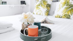 A neatly made bed with white bedding and tropical leaf-patterned pillows, a tray with a mug, glasses, and a flower arrangement.