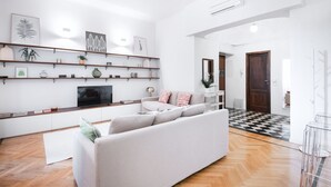 A modern living room with a white sofa, wooden flooring, and a black and white checkered floor mat. Shelves with decorative items and a flat-screen TV are visible.