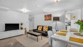 A modern living room with a flat-screen TV, sofa, coffee table, and dining area with a white table and chairs.