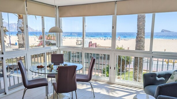 A sunlit room with a glass table and chairs, a sofa, and a view of a beach and mountains. - ESMERALDA sea front apartment (Benidorm)