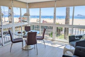 A sunlit room with a glass table and chairs, a sofa, and a view of a beach and mountains. - ESMERALDA sea front apartment (Benidorm)