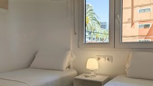 A hotel room with two beds, a nightstand with a lamp, and a window with a view of palm trees and buildings. - ESMERALDA sea front apartment (Benidorm)