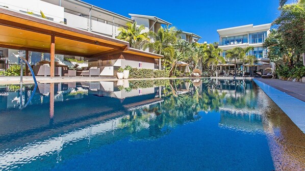 A swimming pool with a wooden deck and lounge chairs, surrounded by modern buildings and tropical vegetation.