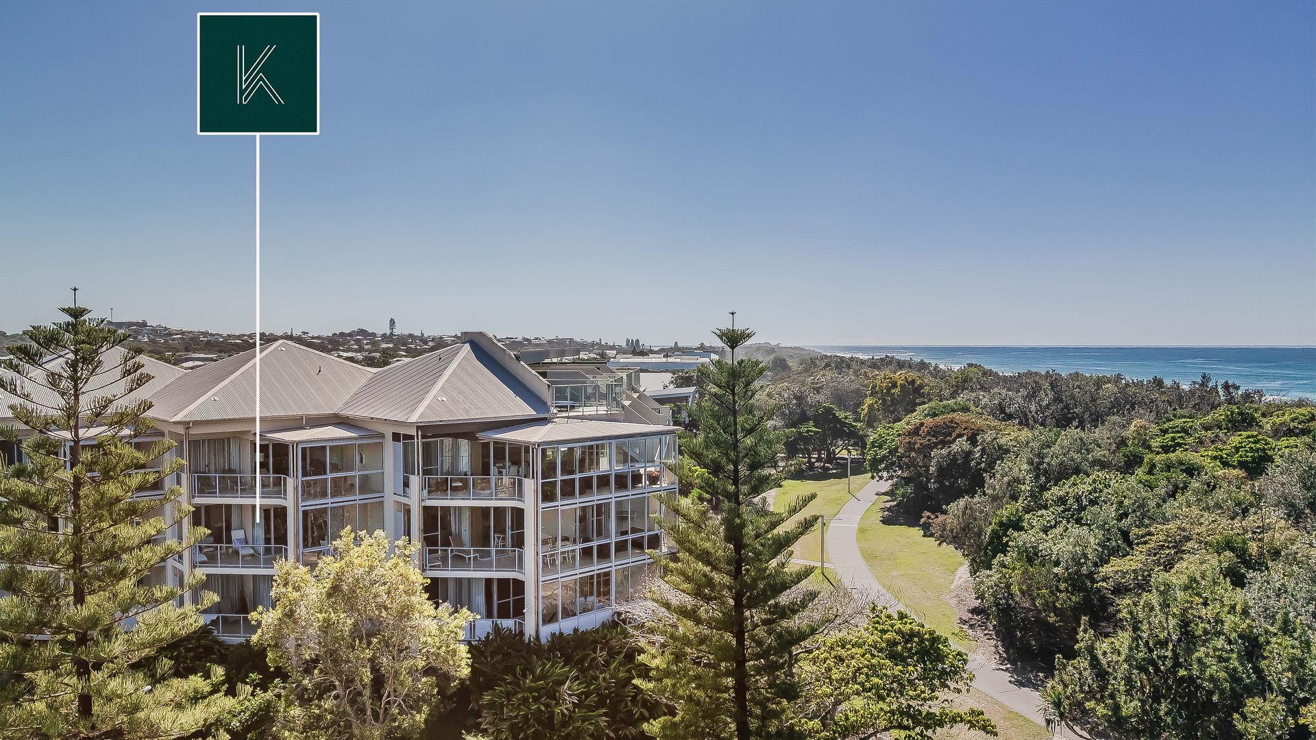 A multi-story building with balconies, surrounded by dense greenery and a clear view of the ocean.