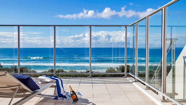 Een balkon met glazen hekwerk met uitzicht op een strand met golven, een ligstoel en een fles champagne.
