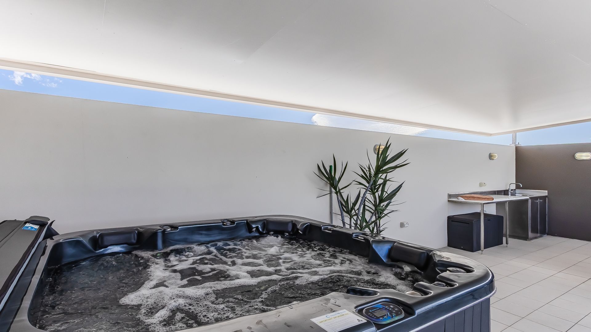Ein Whirlpool mit Bedienfeld, ein kleines Waschbecken und ein Blumentopf in einem modernen Raum mit hohen Decken und großen Fenstern.