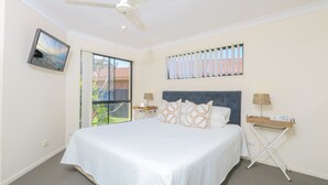 A bedroom with a bed, a ceiling fan, a TV mounted on the wall, a window with blinds, and a small table with a lamp.