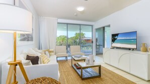 A modern living room with a sofa, coffee table, and TV, featuring a balcony with a view of the beach. - Drift Pool View 21 - Elegant Apartment with Spectacular Lagoon Pool and Tropical Garden Views (Casuarina)