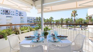 Un balcón con mesa lista para dos personas, sillas de mimbre blanco y vistas a palmeras y edificios.