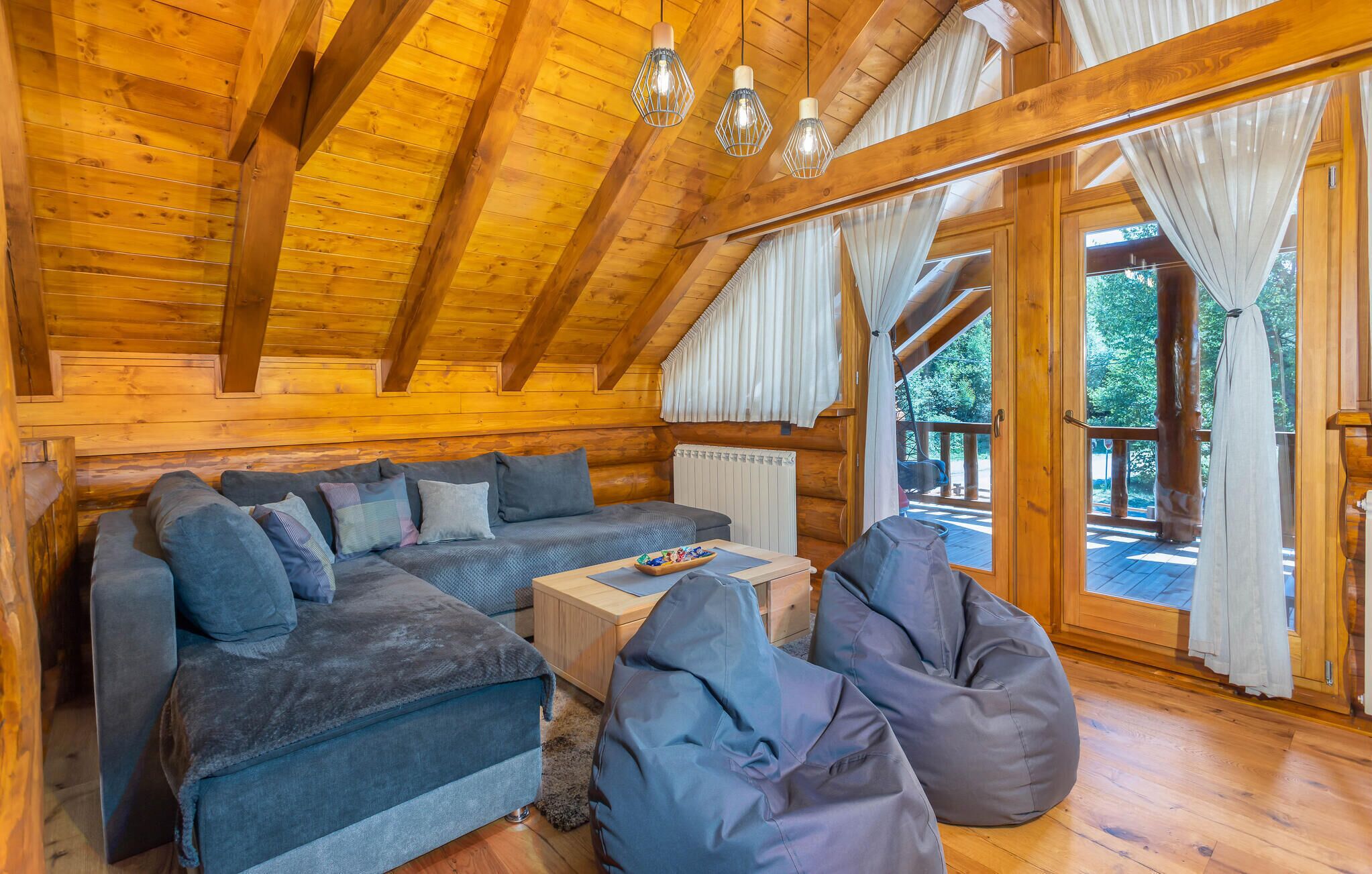 A cozy log cabin interior with a wooden ceiling, a sofa set, bean bags, and a view of the outdoors.