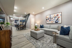 A modern living room with a grey sectional sofa, a dining table with chairs, a staircase, and a kitchen in the background. - Indy Elegance | Minutes to Downtown | Free Parking (Indianapolis)