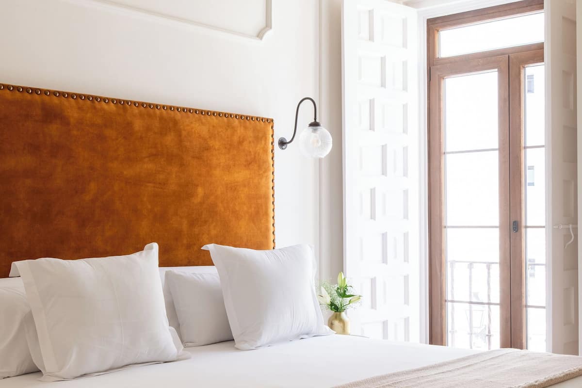 A neatly made bed with white linens and a brown headboard, a bedside lamp, and a view of the outside through an open door.