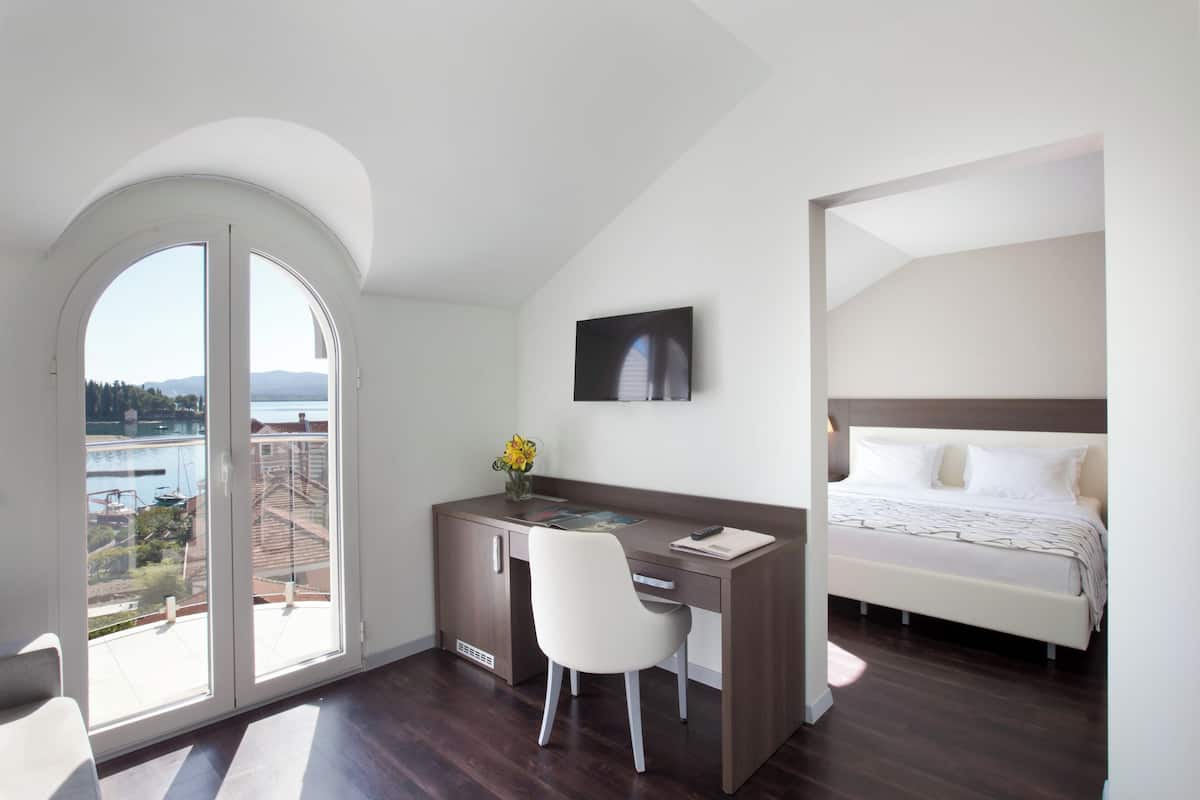 A bedroom with a bed, a desk with a chair, a window with a view of a body of water and buildings, and a painting on the wall.