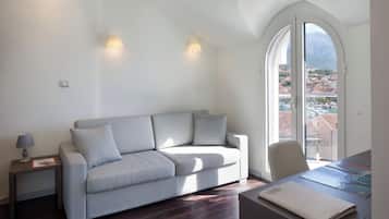 A modern living room with a grey sofa, a small table with a lamp, and a view of a cityscape through large windows.