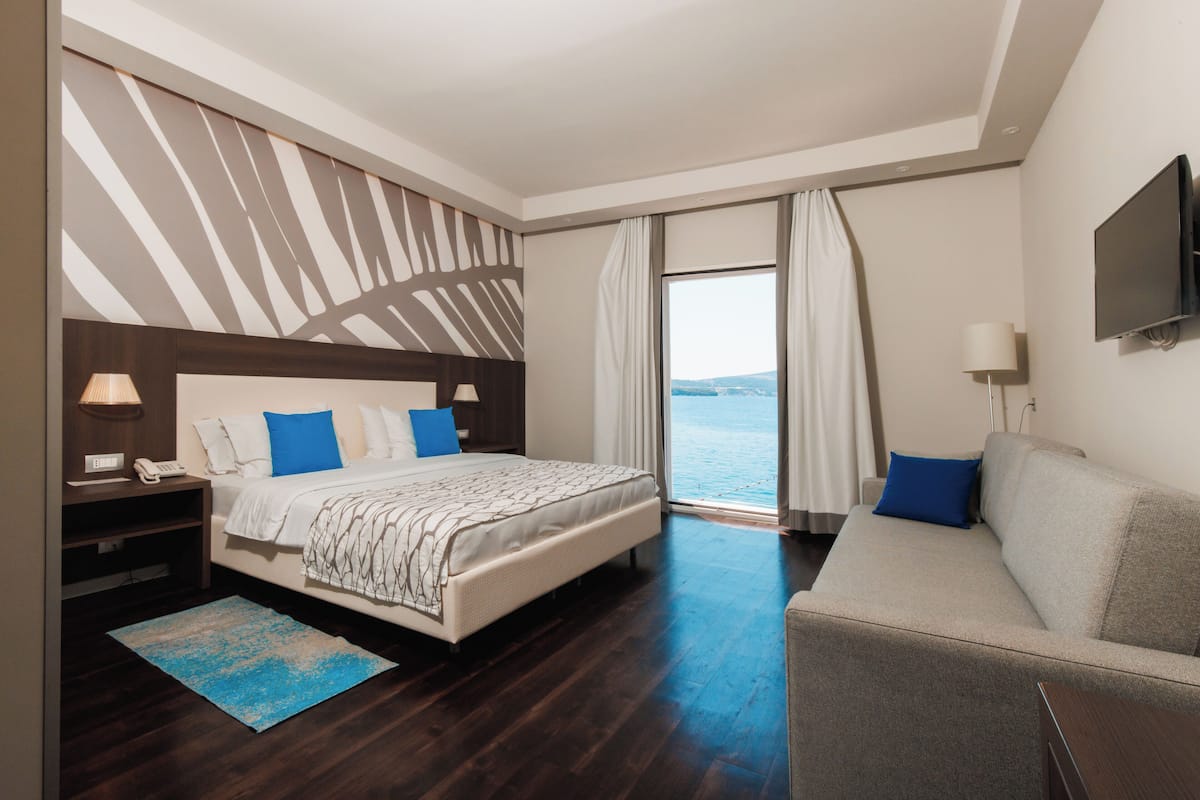 A modern hotel room with a large bed, a sofa, a TV, and a view of the sea.