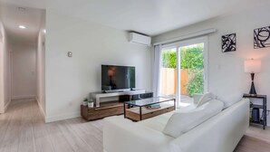 A modern living room with a white sofa, a glass coffee table, a flat-screen TV, and a sliding glass door leading to a garden. - A/C, 3 blocks to beach, dogs, shopping, dining (San Diego)