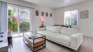 A modern living room with a white sofa, a glass coffee table, and a TV stand. There are abstract wall art pieces and a lamp on a side table. - A/C, 3 blocks to beach, dogs, shopping, dining (San Diego)