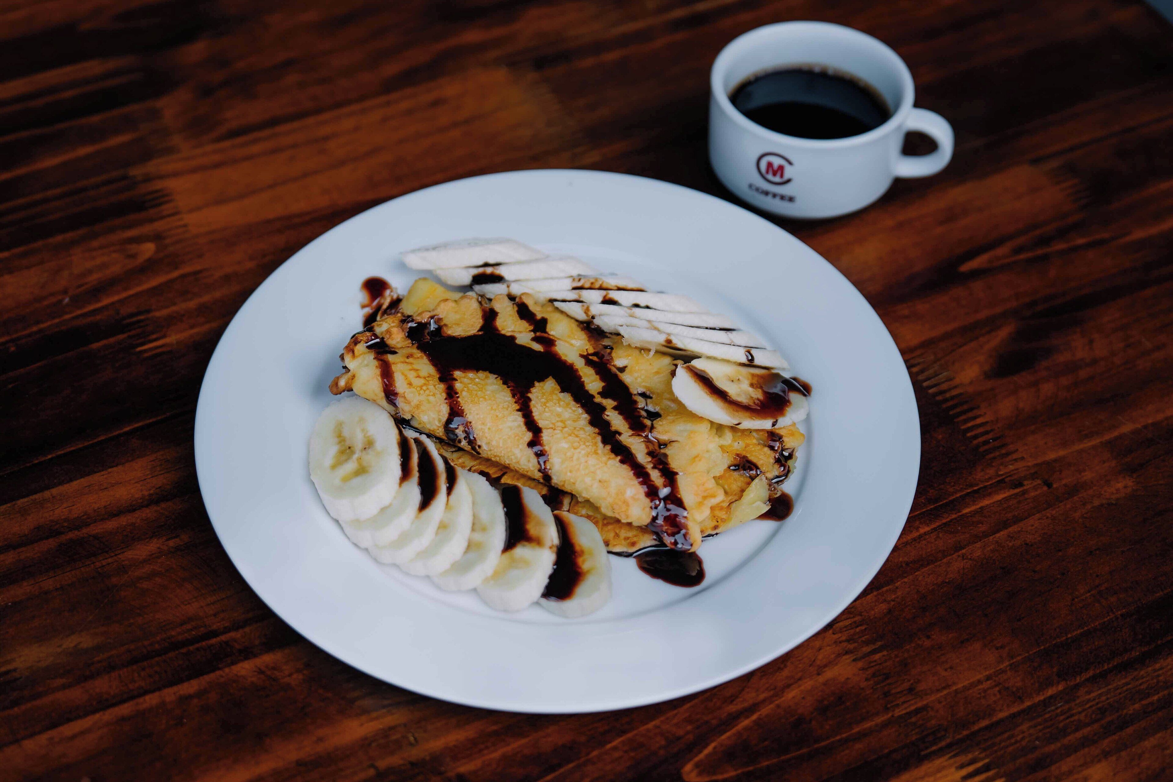 A plate of crepes with chocolate sauce and banana slices on a wooden table.