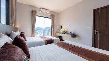 Habitación de hotel con dos camas, un ventanal con cortinas, un equipo de aire acondicionado, una mesita con un florero y una puerta de madera.