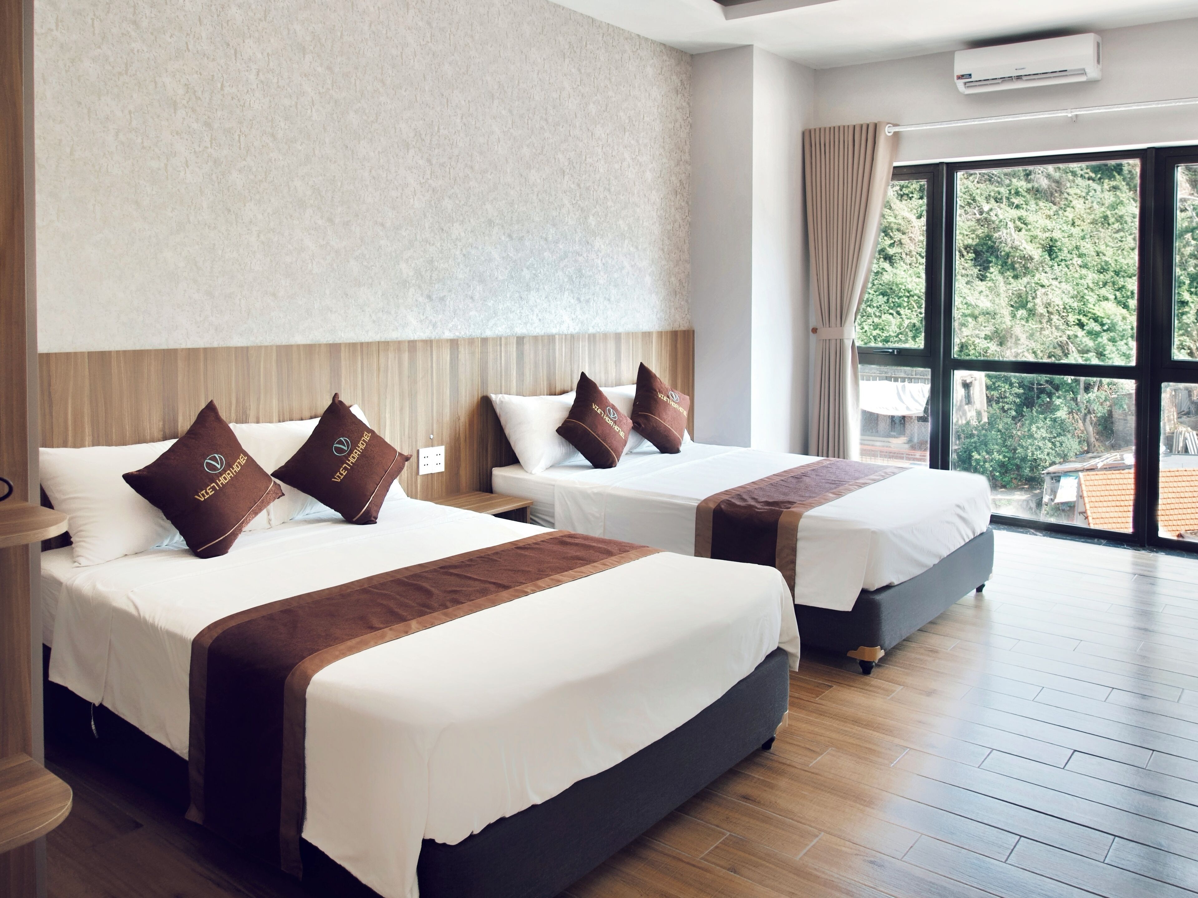 Une chambre d’hôtel avec deux lits, chacun agrémenté de draps blancs et d’oreillers marron, une grande fenêtre donnant sur un paysage verdoyant et un climatiseur fixé au mur.
