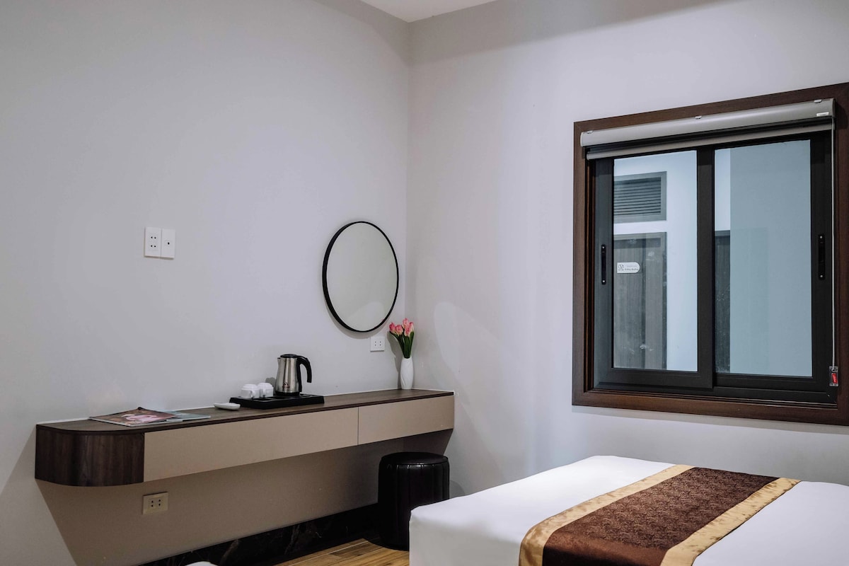 Ein Hotelzimmer mit Bett, Nachttisch mit Lampe, Spiegel, Fenster und kleinem Papierkorb.
