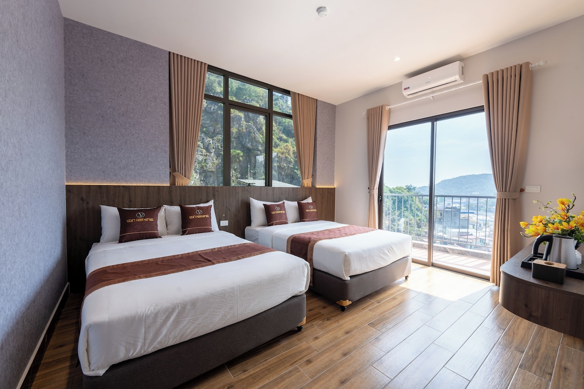 A hotel room with two beds, a large window with curtains, a balcony, and a wooden floor.