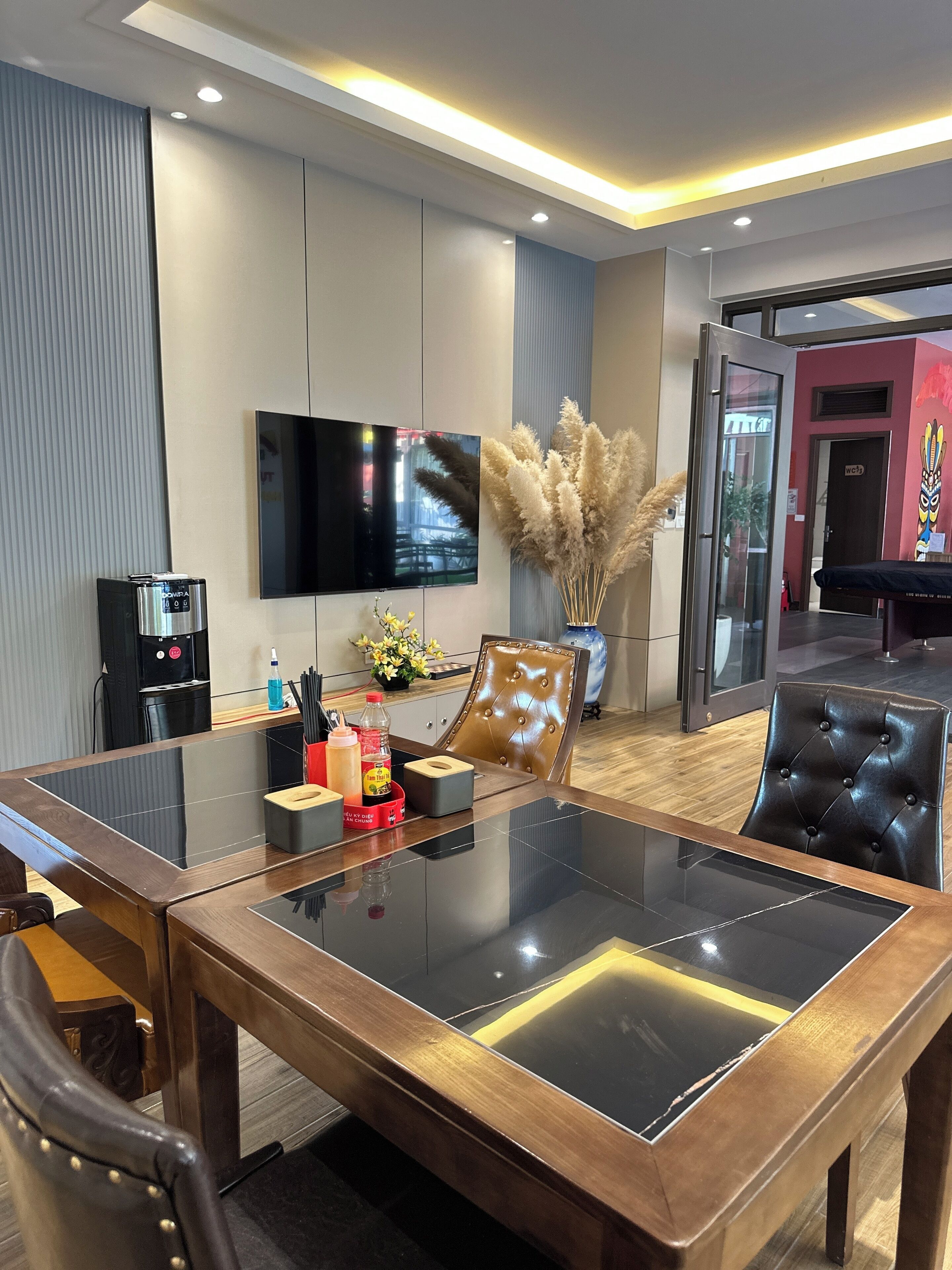 Un espace repas moderne avec une table à plateau en verre, des chaises en cuir, un téléviseur fixé au mur et une machine à café.
