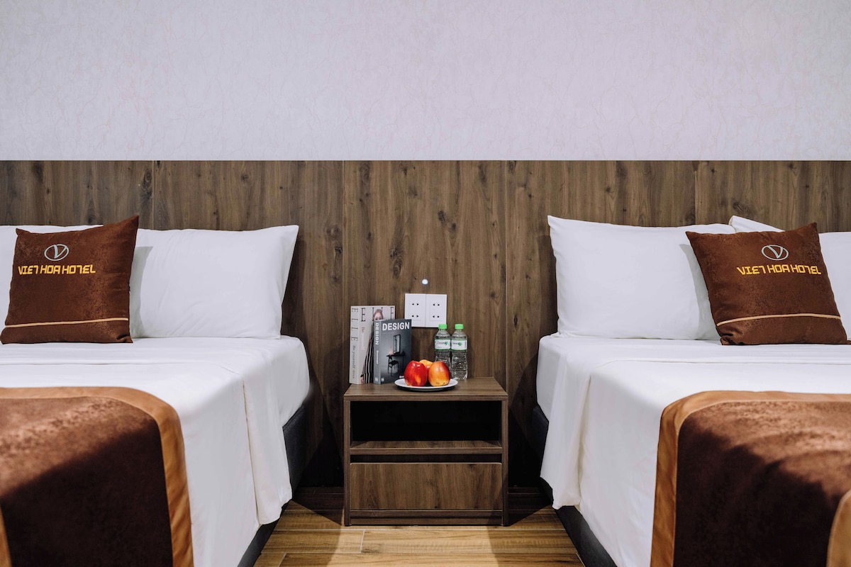 Ein Hotelzimmer mit zwei Betten, jeweils mit einem braunen Kopfkissen und einer weißen Bettdecke. Dazwischen befindet sich ein kleiner Nachttisch aus Holz mit einer Fruchtschale und zwei Wasserflaschen.