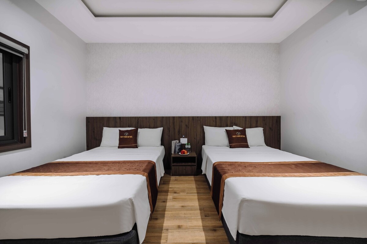 Een hotelkamer met twee bedden, een houten hoofdbord, een klein tafeltje met een schaal fruit en een raam aan de linkerkant.