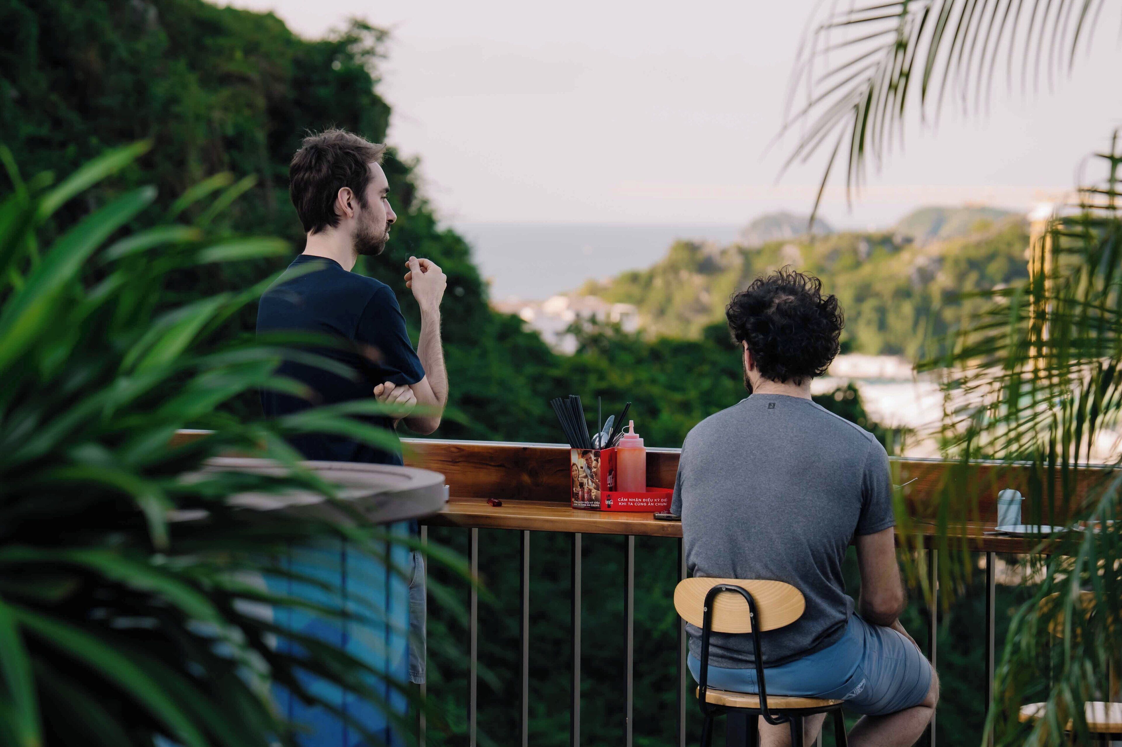 Dos personas sentadas en una mesa de madera con vista al océano y follaje.