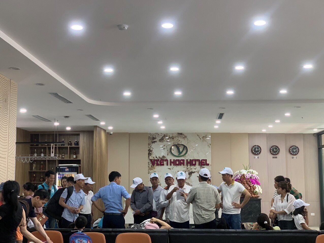 A group of people in a hotel lobby with a reception desk, a clock, and a wall with the hotel's name.