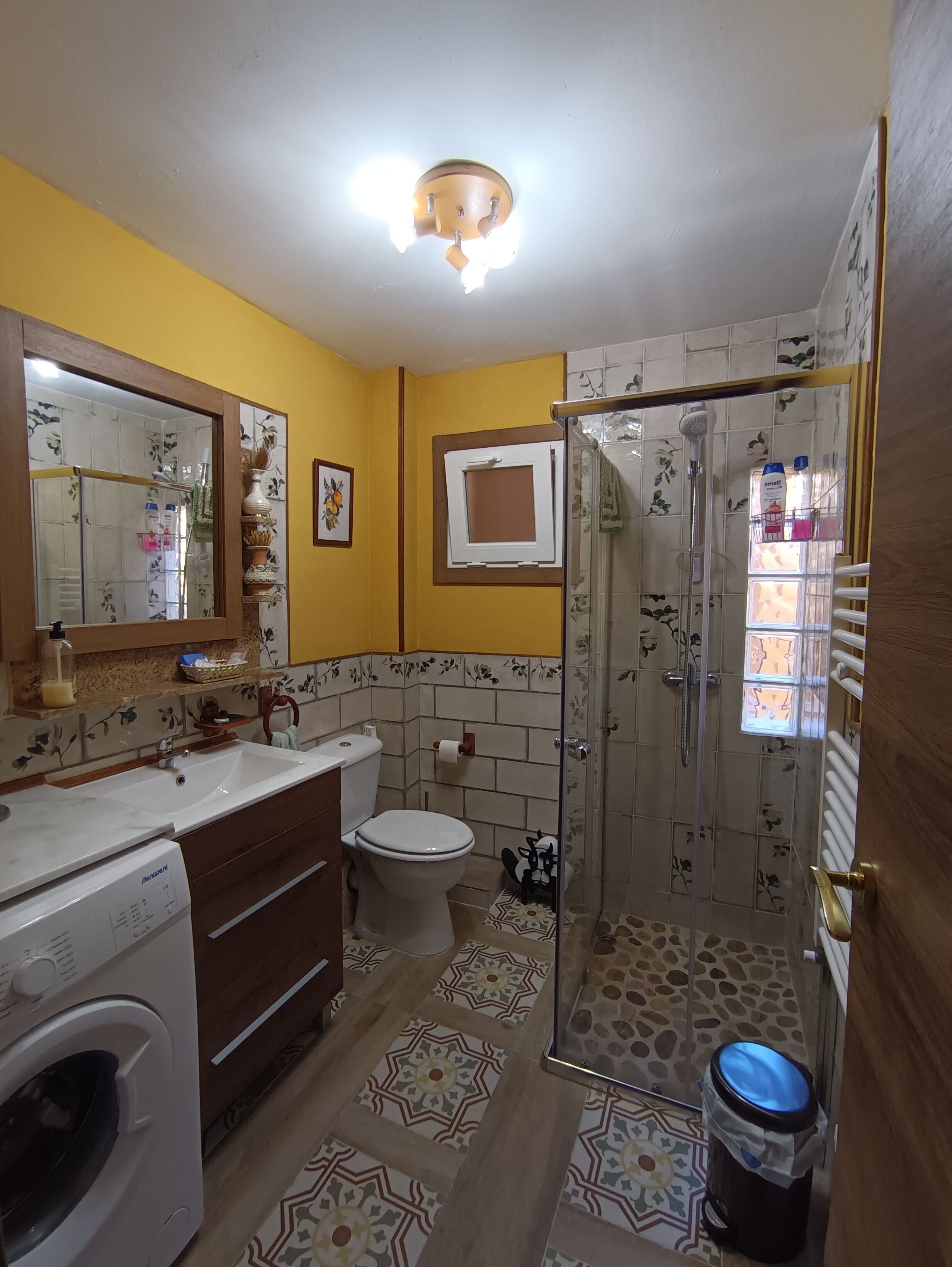 Un baño con una pared amarilla, un lavabo blanco, un inodoro, una ducha con puerta de cristal y una lavadora.