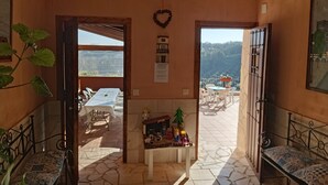 Une pièce avec un sol en pierre, une petite table recouverte d’une nappe blanche, un banc et une vue sur une terrasse extérieure avec des chaises et une table.