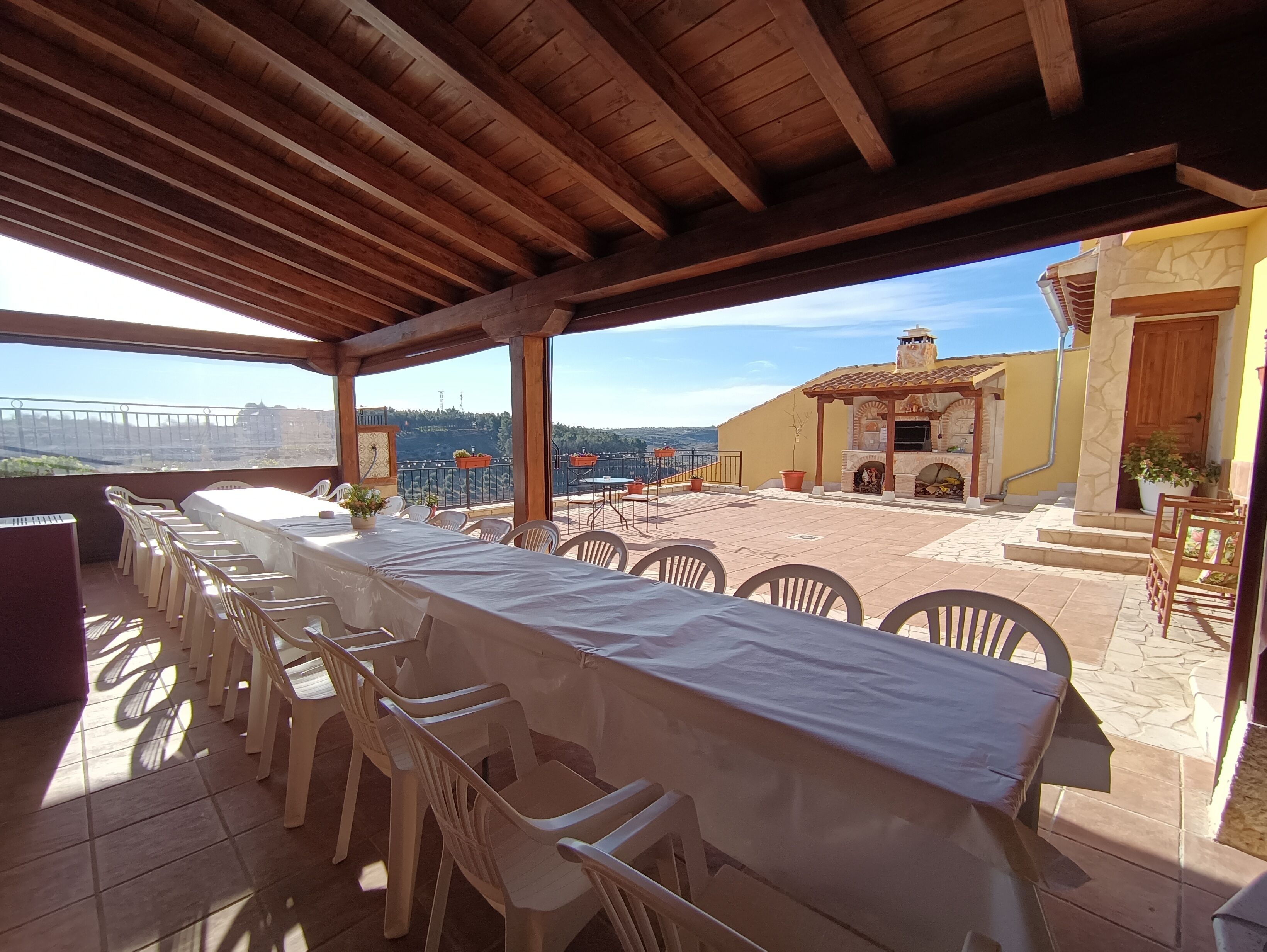 An outdoor patio with a long table and chairs, a covered area with a wooden ceiling, and a view of a building with a stone facade.