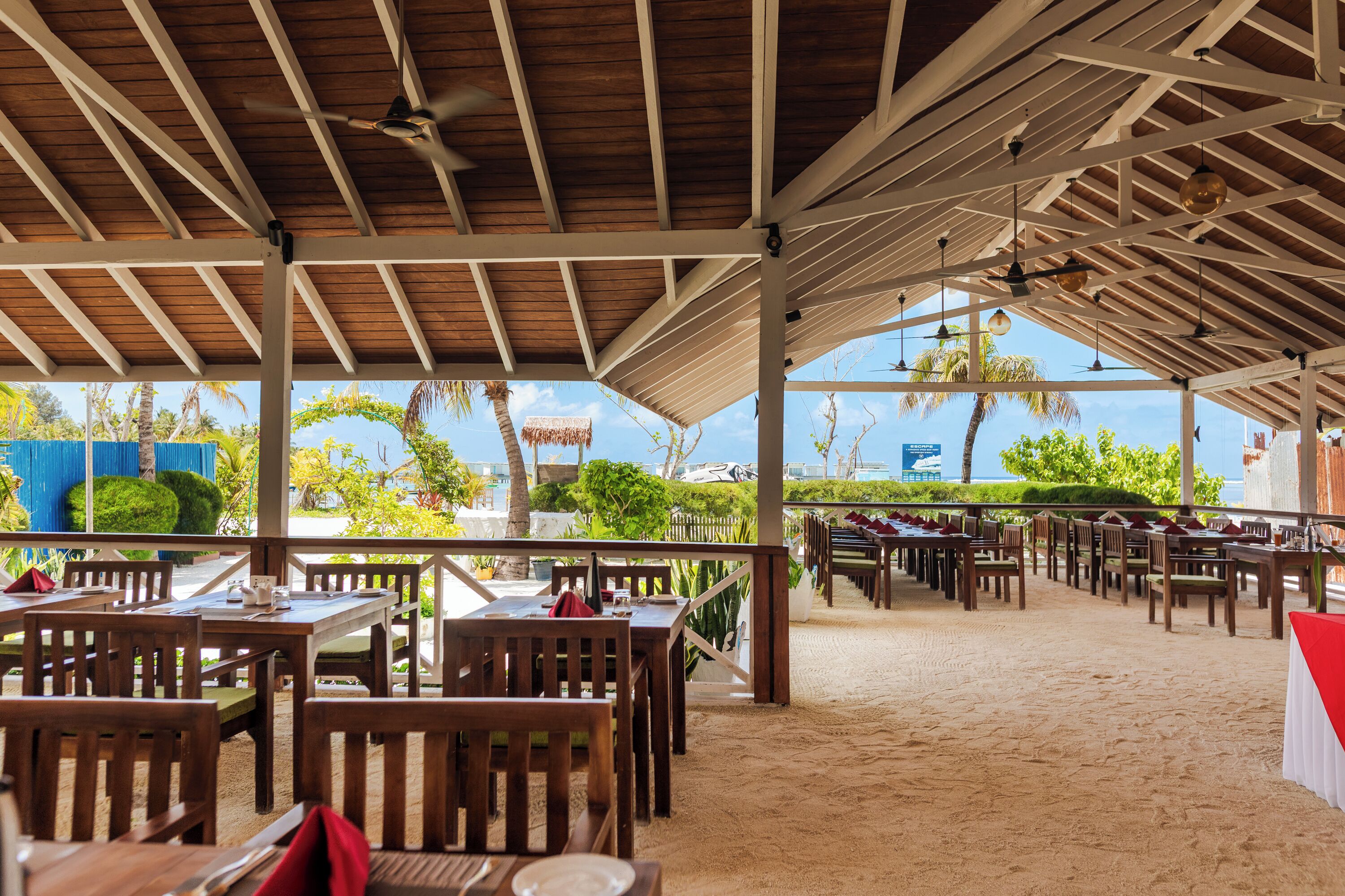 Un restaurante al aire libre con mesas y sillas de madera, suelo de arena y vistas al mar y palmeras.