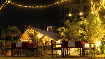 A beachfront dining area with tables set for guests, palm trees, and string lights.