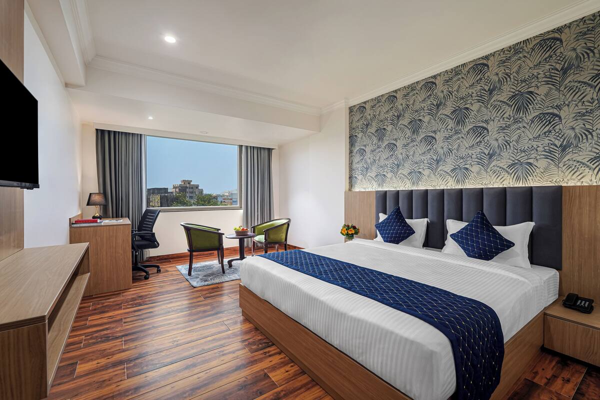 A hotel room with a large bed, a desk, a chair, a small table with a lamp, and a view of the city through large windows.